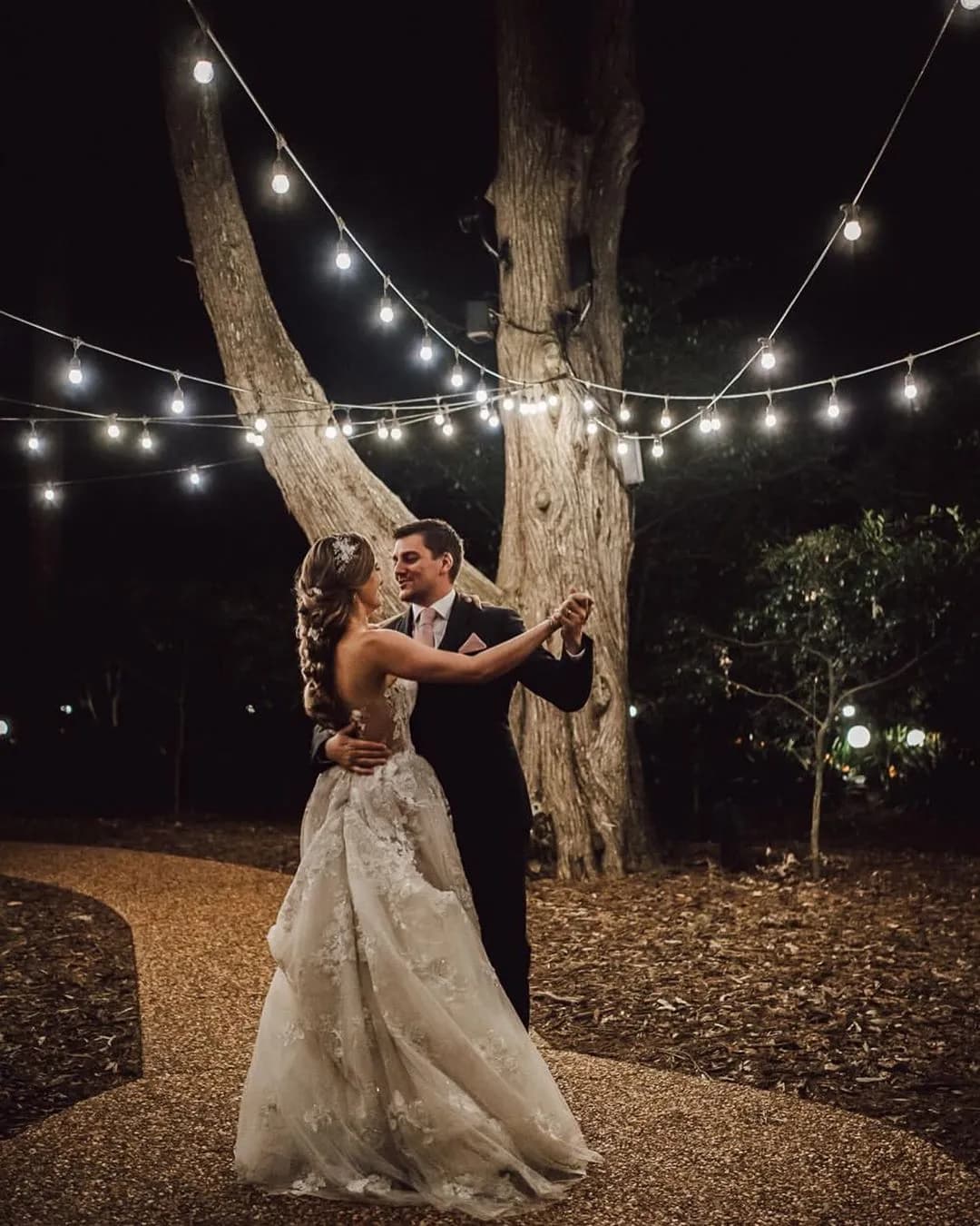 A bride and groom dance under string lights in a nighttime outdoor setting. The bride wears a white lace gown, and the groom is in a dark suit. They are surrounded by trees, creating a romantic atmosphere.