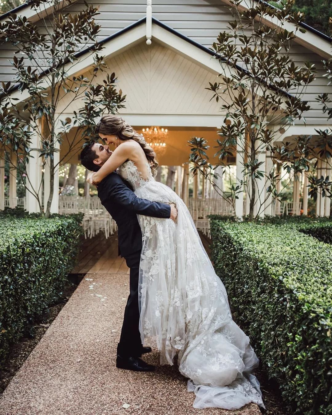 A groom lifts his bride in a joyous embrace in front of an elegant, outdoor wedding chapel. The bride's white gown cascades down as she leans back, smiling at him. The path is lined with manicured hedges, and the chapel is adorned with delicate lights.