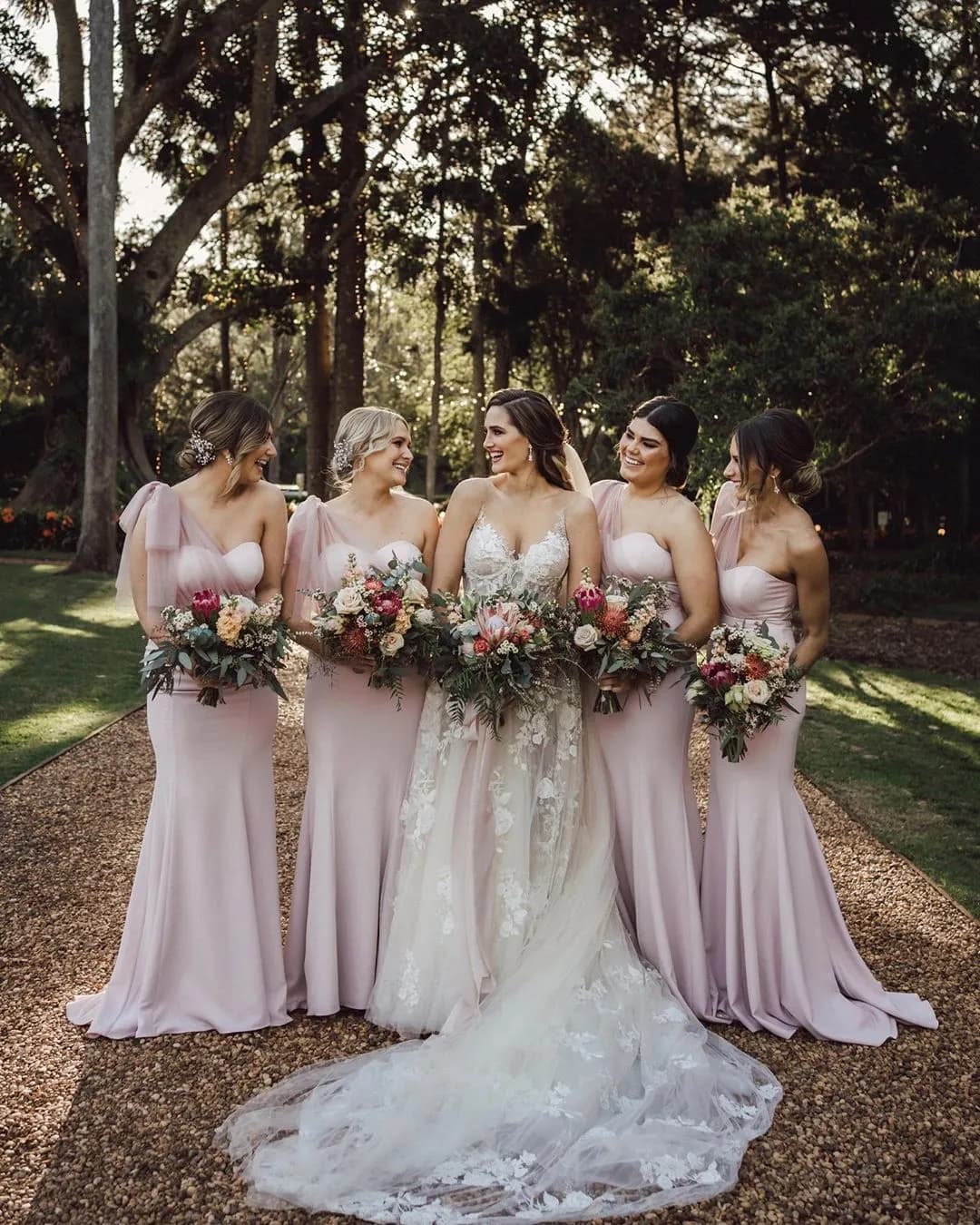 A bride in a white wedding dress stands outdoors with four bridesmaids dressed in matching light pink gowns. Each holds a bouquet of flowers. They are all smiling and standing on a garden path lined with trees and greenery in the background.