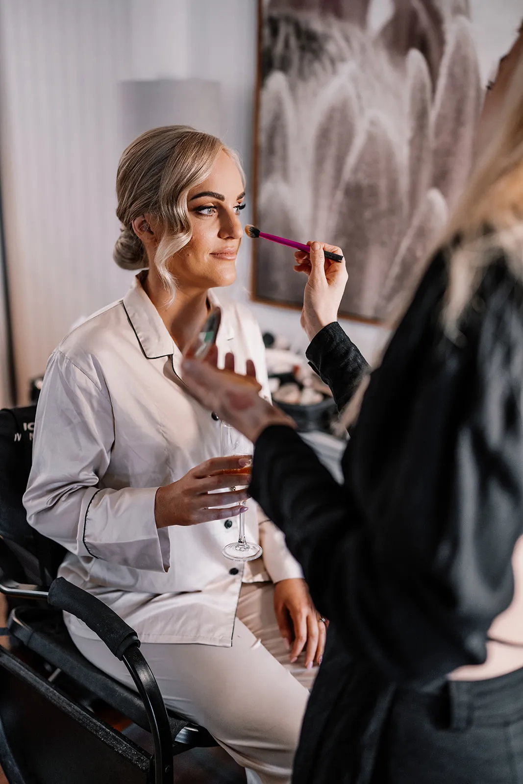 A woman with blonde hair is seated in a chair, wearing a white button-up shirt, and holding a glass. Another person is applying makeup to her face using a brush. The setting appears to be indoors with a blurred background featuring a framed picture.