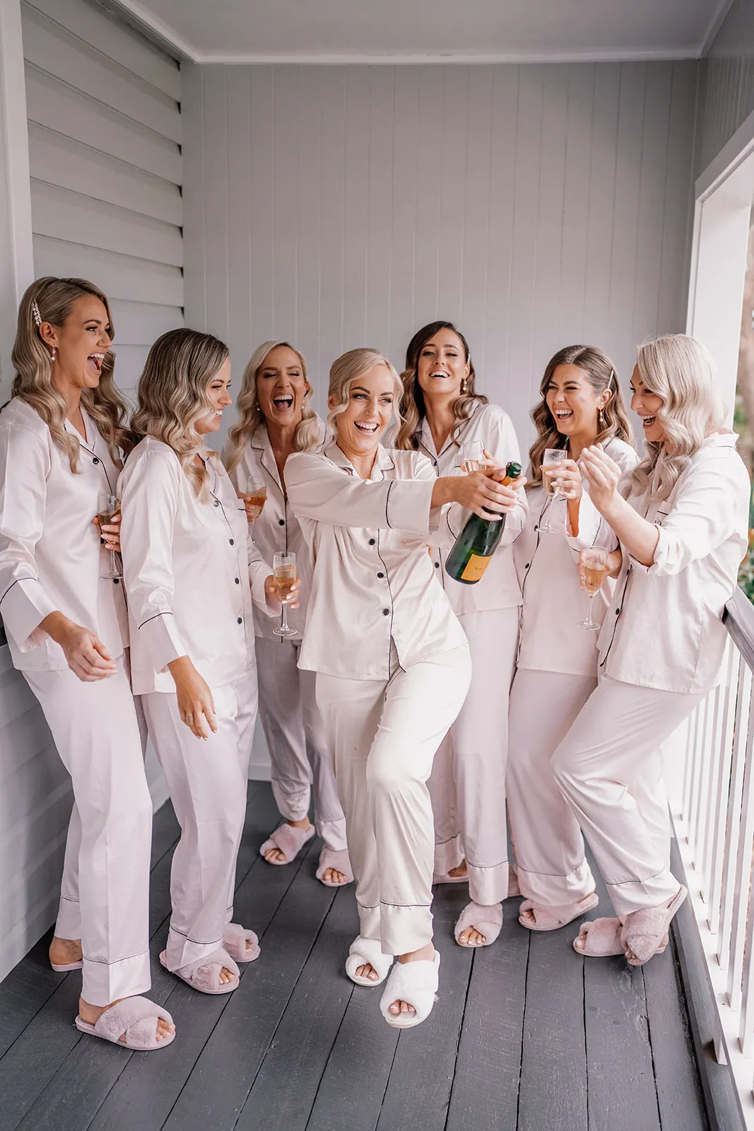 A group of seven women in matching light pink pajamas and slippers stand on a porch, smiling and laughing. One woman in the center pops a bottle of champagne while the others, holding glasses of bubbly, look on happily.