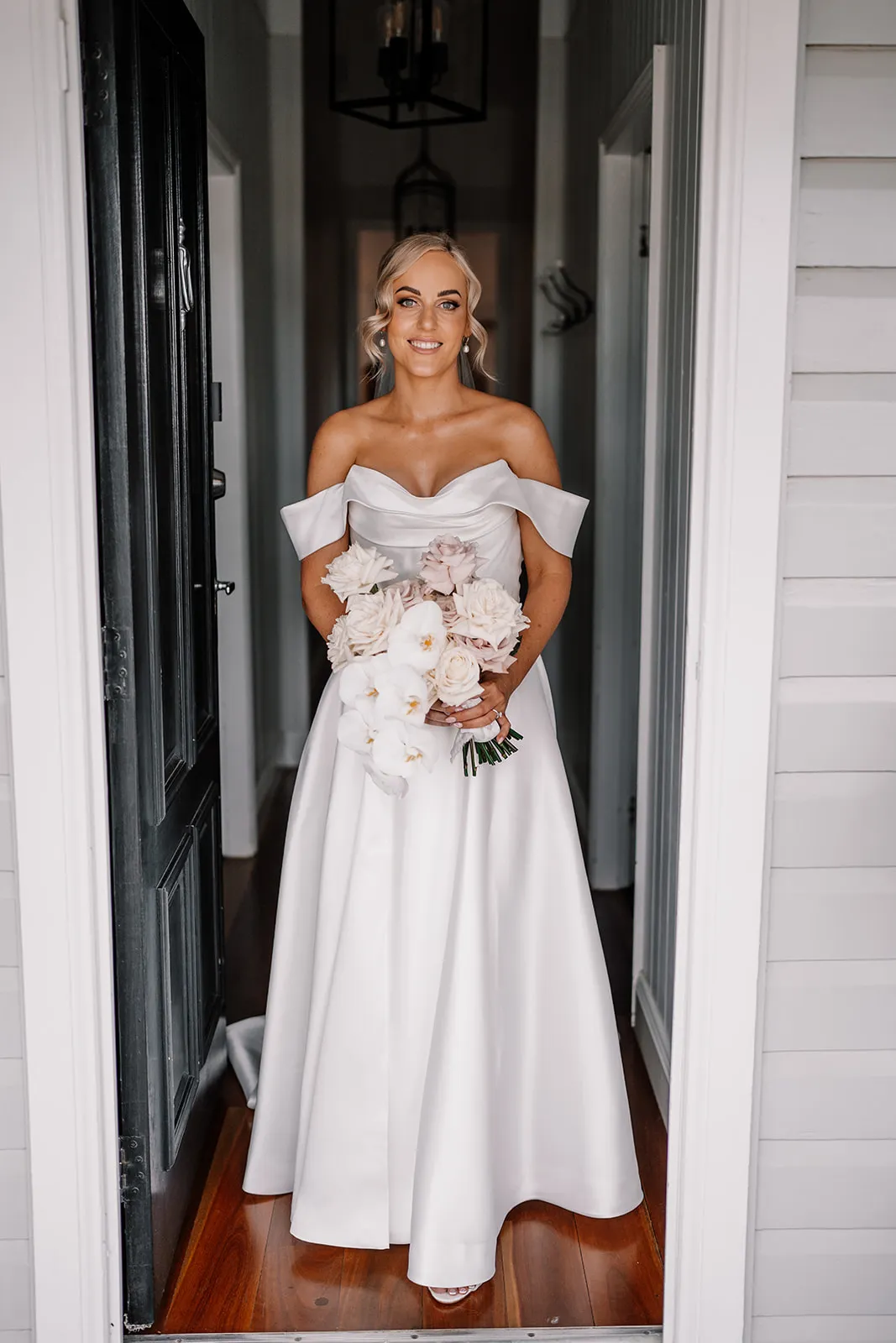 A bride stands in a doorway, smiling. She wears an off-the-shoulder, white satin wedding dress and holds a bouquet of white and blush pink flowers. The background shows a narrow hallway with dark wooden floors and white walls.