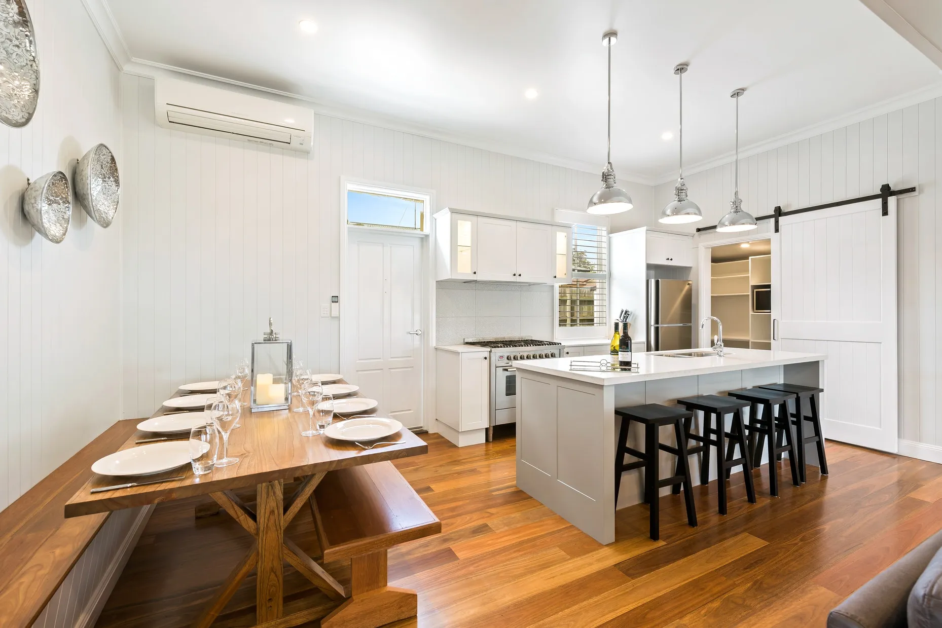 A modern kitchen and dining area with a wooden floor and white walls. The kitchen features a central island with bar stools, stainless steel appliances, and pendant lights. The dining area has a wooden table with bench seating. Decorative plates adorn the wall.