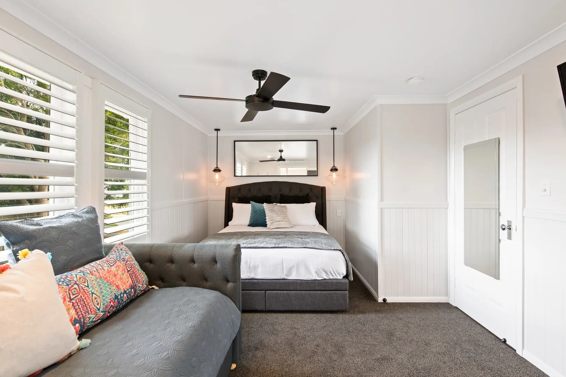 A modern bedroom features a bed with a gray upholstered headboard, white linens, and decorative pillows. There's a tufted gray bench with colorful cushions on the left. The room has large windows with white blinds, two pendant lights, and a ceiling fan.