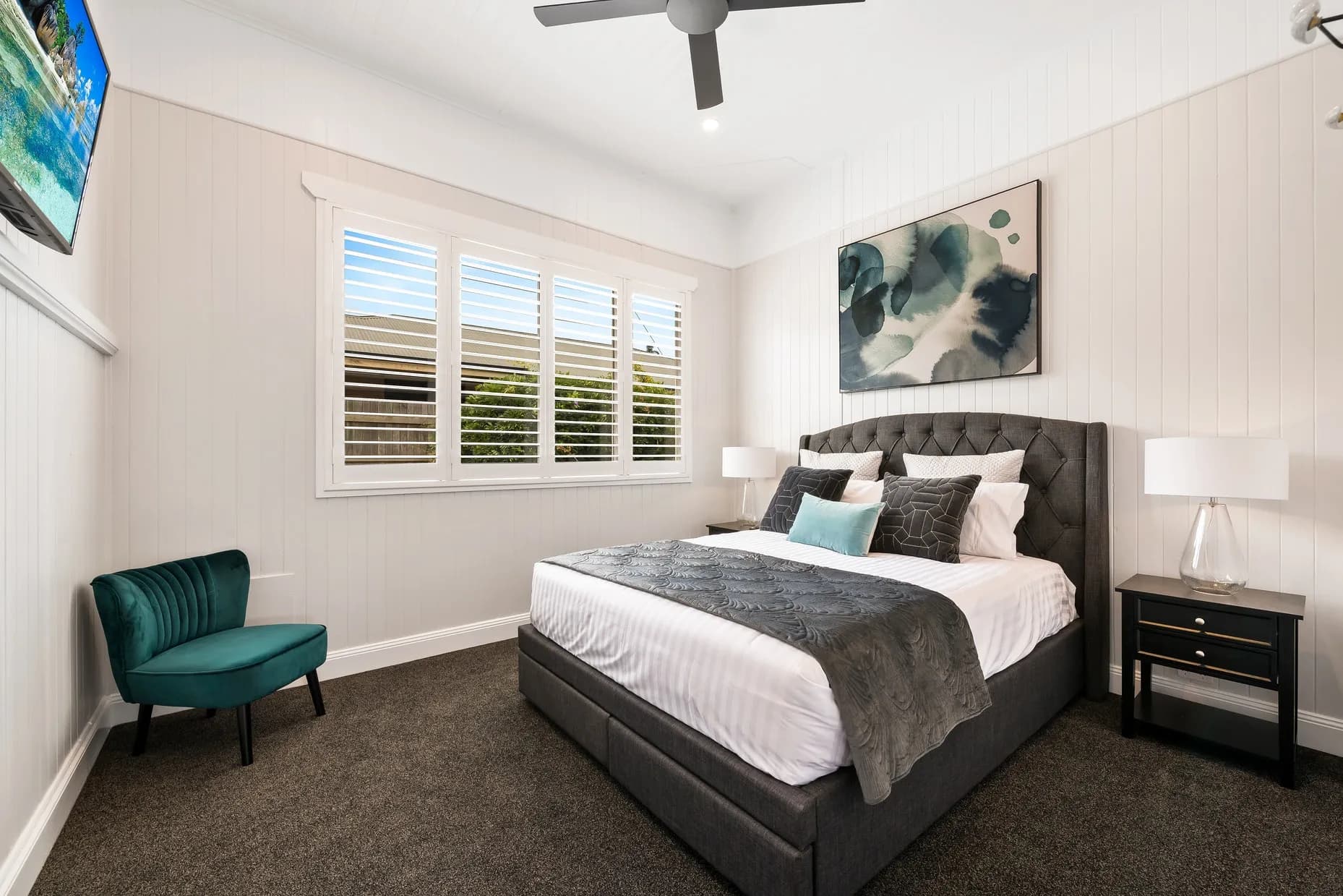 A modern bedroom with white paneled walls, grey carpet, and a large window with white shutters. The bed has a grey upholstered headboard, white linens, and grey and teal pillows. There are two nightstands with lamps, a green chair, and a ceiling fan. A painting and wall-mounted TV are on the walls.