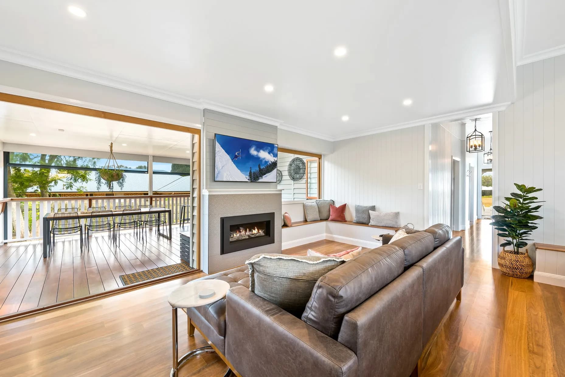 Modern living room with a leather sofa facing a wall-mounted TV above a fireplace. The room extends to a wooden deck with a dining table, accessible through large sliding glass doors. The space is well-lit with recessed lighting and natural light.