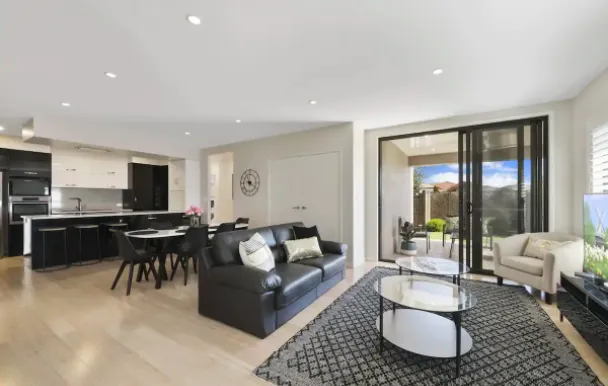 A modern, open-concept living room and kitchen area with large sliding glass doors leading to the backyard. The room is furnished with a black leather sofa, armchairs, a coffee table, a dining table with chairs, and a wall-mounted TV. The decor is minimalistic and contemporary.