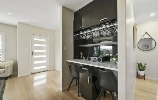 A modern room featuring a small bar with a mirrored backsplash, wine glasses hanging overhead, and two black bar stools. Adjacent is a light hardwood floor leading to a front door with horizontal glass panel inserts. A cozy living area is partially visible on the left.
