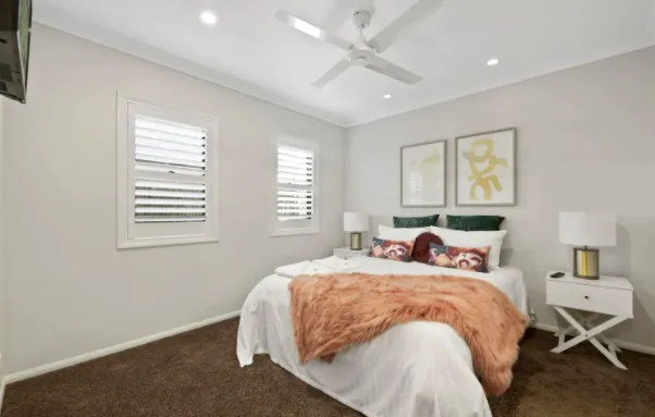 A cozy bedroom features a double bed with white bedding, a brown throw blanket, and decorative pillows. There are two bedside tables with lamps, two framed pictures on the wall, a ceiling fan, and two shuttered windows allowing natural light into the room.