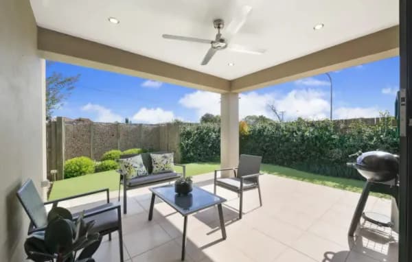 A covered outdoor patio with a ceiling fan, a glass-top table, and four modern chairs. The floor is tiled, and a potted plant sits beside the entrance. Adjacent to the seating area is a barbecue grill. The patio overlooks a well-manicured lawn and greenery.