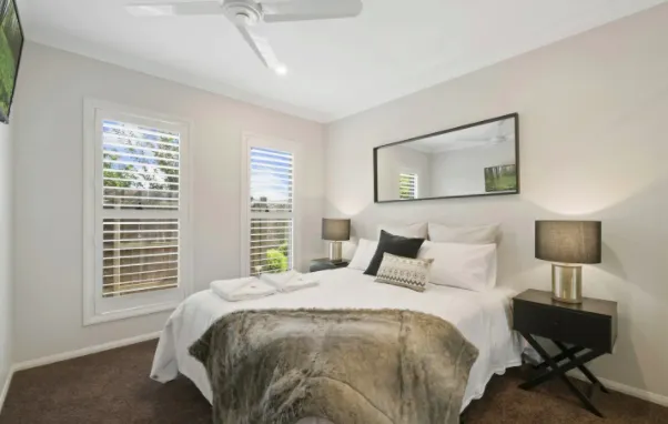 A cozy bedroom with a double bed adorned with white linens and a large fur throw blanket. Two bedside tables with lamps flank the bed. A wide mirror is mounted above the bed. A ceiling fan is overhead, and natural light enters through two shuttered windows.