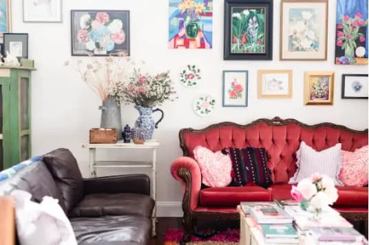 A cozy living room with a red tufted sofa, a brown leather couch, and a small white table holding a blue floral vase. The walls are decorated with an assortment of framed colorful artwork. A coffee table is adorned with books, magazines, and flowers in the foreground.