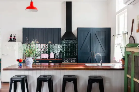 A modern kitchen featuring dark cabinets, a black range hood, a wooden countertop with four black stools, and a farmhouse-style sink. The backsplash is glossy green tile, a red pendant light hangs from the ceiling, and various plants and decor adorn the space.