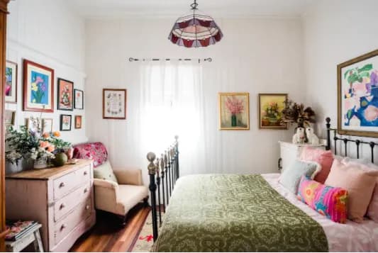 A cozy bedroom with a vintage touch features a black metal bed frame, colorful quilts, and plump pillows. A pink dresser with flowers in a vase, an upholstered chair, and framed artwork on the white walls add a vibrant and inviting feel to the room.