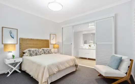 A bright and modern bedroom with a beige bed, two bedside tables with lamps, and two bird-themed framed pictures above the bed. There is a cushioned chair in the corner, and a sliding door that opens into a bathroom with a white vanity and mirror.