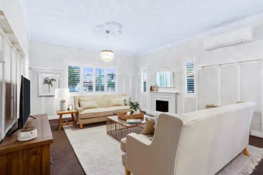 A well-lit living room with white walls featuring a comfortable seating area that includes a beige sofa, armchair, and wooden coffee table. There is a fireplace with a mirror above it, a wall-mounted TV and a modern chandelier hanging from the ceiling.