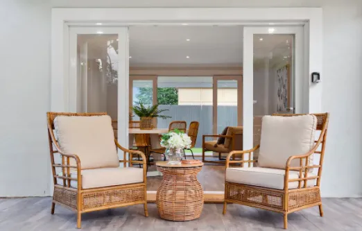 A cozy patio setup with two beige cushioned rattan chairs and a small wicker table topped with a vase of white flowers. The patio overlooks a modern interior through large sliding glass doors, revealing a dining table and chairs.