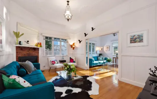 A brightly lit living room with white walls features a teal sofa, a glass coffee table on a black and white cowhide rug, and a fireplace with a mantel. Wall art and flying bird ornaments decorate the space, leading to another room with additional seating and decor.
