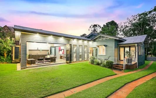 A modern, single-story contemporary house with a green lawn and a brick pathway leading to the entrance. The home features large glass sliding doors, an outdoor sitting area, and is illuminated by exterior lights in the evening with a multi-colored sky in the background.