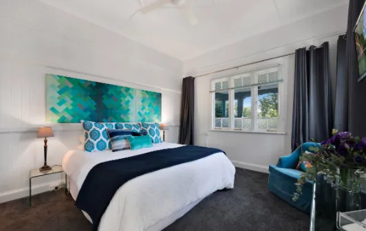 A well-lit bedroom featuring a large bed with white linens, blue throw pillows, and a dark blue blanket. Above the bed is a vibrant abstract painting in shades of blue and green. The room includes a window with dark curtains and a blue armchair next to a small side table.