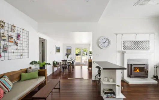 A modern living area with white walls and wooden flooring features a lounge with a bench and cushions, a fireplace with a mantel, a wall clock, and a dining area in the background with a table and chairs. A grid display of photos is on the wall, and large windows let in natural light.