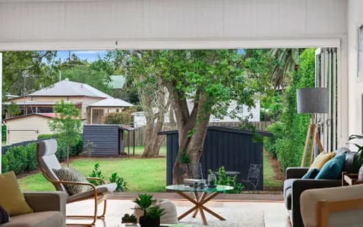 A cozy living room with a large open sliding door leading to a lush green backyard. The room features comfortable seating, including a chair and a sofa, a stylish floor lamp, and a coffee table. The backyard has a small shed and is surrounded by trees and greenery.