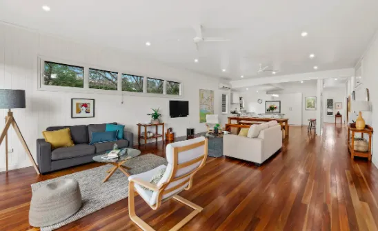 A spacious living room with wooden flooring, featuring a gray couch, a mid-century modern armchair, and a light-colored sofa. The room is well-lit with several windows near the ceiling. A dining area and kitchen are seen in the background. Stylish decorations adorn the space.