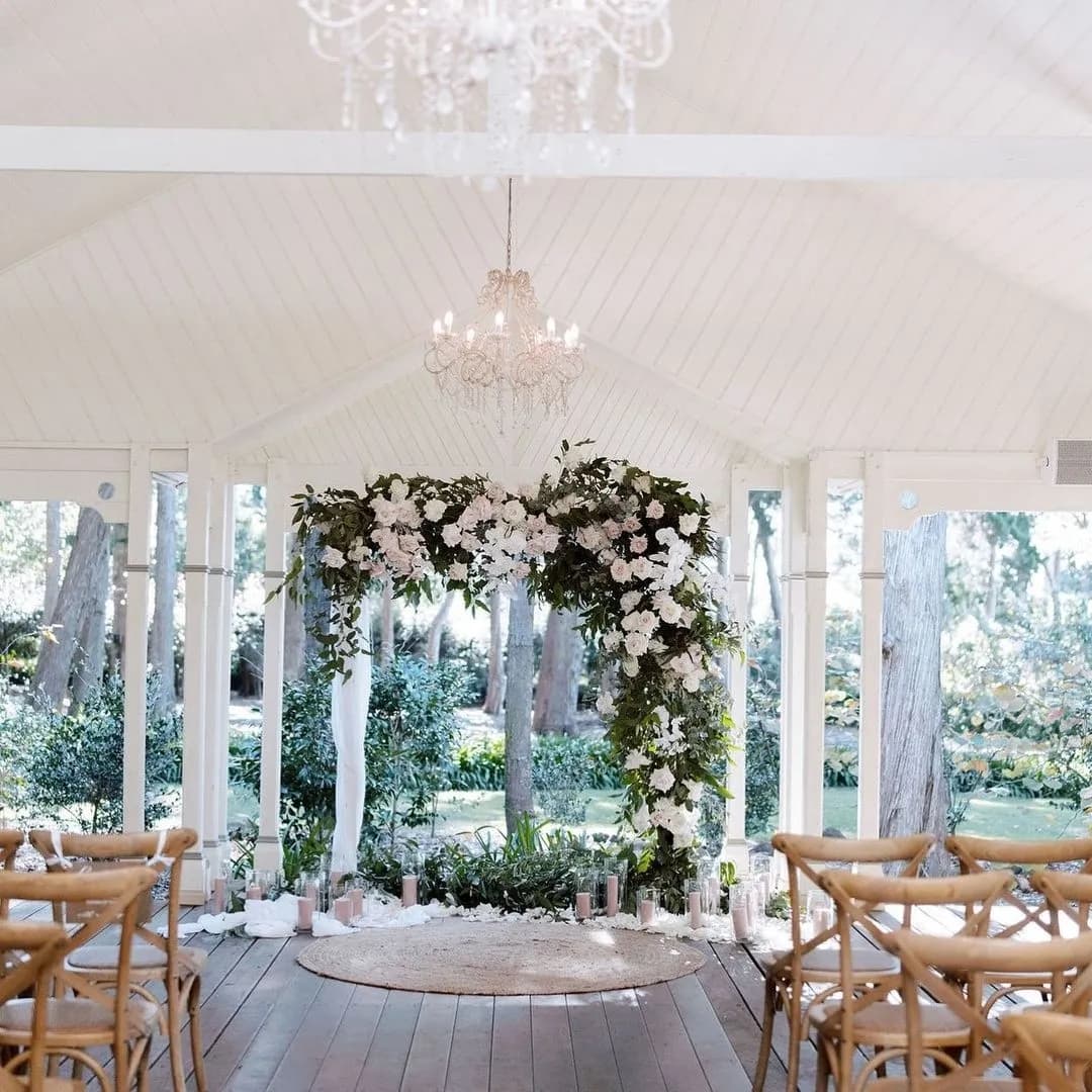 An elegant wedding ceremony setup featuring a lush floral arch adorned with white flowers and greenery. Wooden chairs are arranged neatly on either side of the aisle. The ceiling is draped with white fabric and a chandelier hangs above, adding a touch of sophistication.