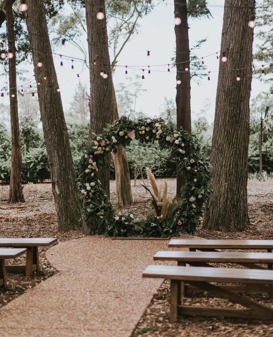 A serene outdoor wedding setup in a forested area, featuring a large, circular floral arch adorned with greenery and flowers. Wooden benches are placed on either side of a gravel aisle, and string lights are hung between trees, creating a romantic atmosphere.