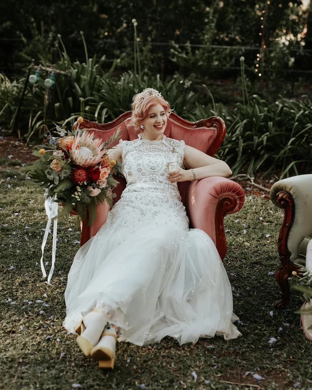 A bride in a white, embellished wedding dress sits on a red vintage armchair outdoors, smiling and holding a glass. She has a large bouquet of colorful flowers in her lap, and there is greenery in the background, creating a serene and festive setting.