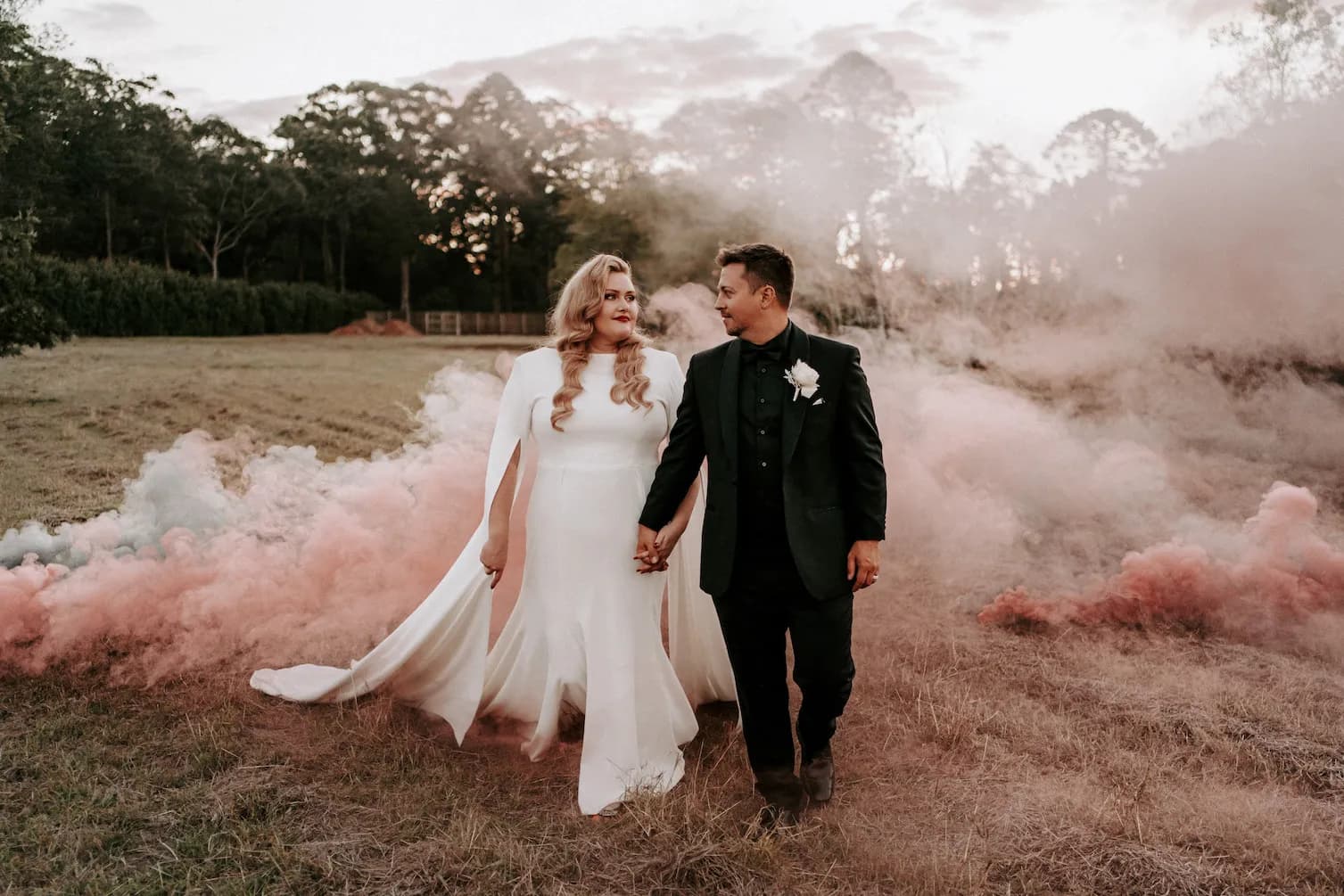 A bride and groom hold hands and smile at each other while walking through a grassy field with a backdrop of smoky pink and white clouds. The bride wears a flowing white dress, and the groom is dressed in a black suit with a white boutonniere. Trees are in the background.