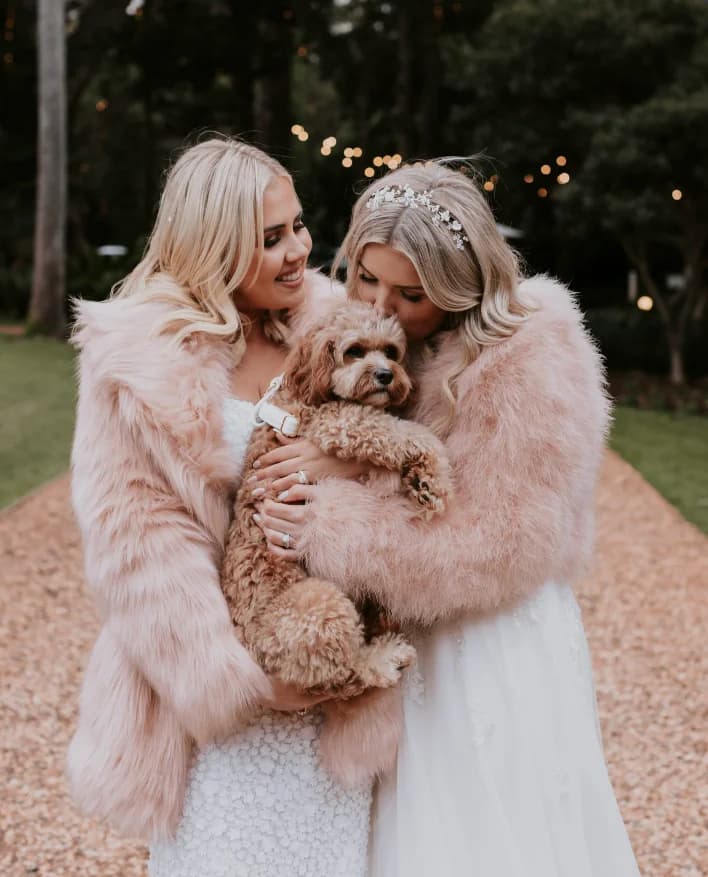 Two women in formal dresses and pink faux fur jackets smiling together outdoors, holding a small brown fluffy dog. One woman has a jeweled headband and is kissing the dog. Trees and string lights are visible in the background.